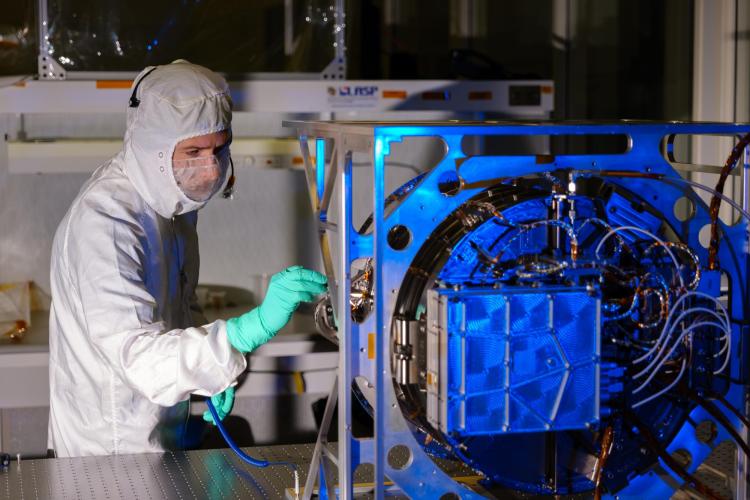 Engineer in protective gear inspects a metal space instrument with wires coming out of its back